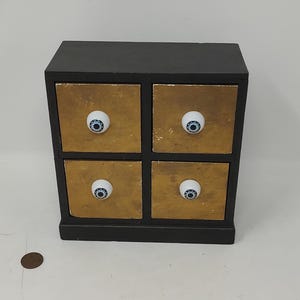 May include: A small, black wooden cabinet with four drawers. The drawer fronts are gold-colored with a textured appearance. Each drawer has a white knob with a blue and black eye design. A unique storage solution.