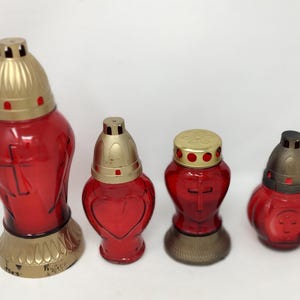 May include: Four red glass memorial candle holders with gold-colored lids and bases. The candle holders have cross and heart designs. The candle holders are of varying sizes and shapes.