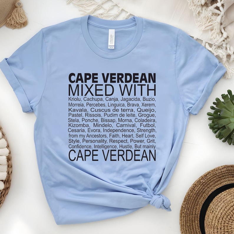 Może przedstawiać: Jasno błękitny t-shirt z czarnym tekstem, kt&oacute;ry brzmi "Cape Verdean Mixed With" i wymienia r&oacute;żne słowa i frazy związane z kulturą Wysp Zielonego Przylądka. Tekst zawiera "Kriolu, Cachupa, Canja, Jagacida, Buzio, Morreia, Percebes, Linguica, Brava, Xerem, Kavala, Cuscus de terra, Queijo, Pastel, Rissois, Pudim de leite, Grogue, Stela, Ponche, Bissap, Morna, Coladeira, Kizomba, Mindelo, Karnawał, Piłka nożna, Cesaria, Evora, Niepodległość, Siła, od moich przodk&oacute;w, Wiara, Serce, Miłość do siebie, Styl, Osobowość, Szacunek, Moc, Odwaga, Zaufanie, Inteligencja, Praca, Ale przede wszystkim Cape Verdean."