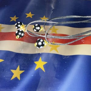 Op de afbeelding: Verschillende kettingen met zwart-witte polkadot kralen en zilverkleurige koorden worden getoond. De kralen hebben een goudkleurig accent. De achtergrond toont een vlag met blauwe, rode, witte en gele kleuren.