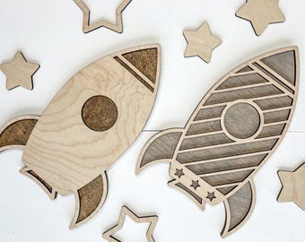 May include: Two wooden rocket ship cutouts with star accents. The rockets are light brown with darker brown accents and circular windows. Several wooden star cutouts are scattered around the rockets, creating a playful, space-themed design.