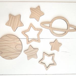 May include: Wooden cutouts of celestial bodies, including stars and planets. The set features solid and open star shapes, a planet with rings, and a circular planet with etched details. The cutouts are made of light-colored wood.