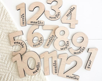 May include: Wooden milestone markers with numbers 1-12 and the word "months" or "year" in a black script font. The numbers are in a sans-serif font and are arranged on a white surface with a cream-colored blanket.