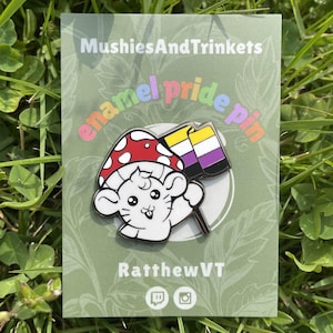 Puede incluir: Pin de orgullo de esmalte con un personaje de hongo de dibujos animados blanco con una gorra de lunares rojos y blancos, sosteniendo una bandera de orgullo no binario. El pin está en una tarjeta verde con el texto "MushiesAndTrinkets", "enamel pride pin" y "RatthewVT".