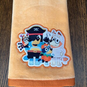 May include: A soft peach-colored hand towel featuring a central embroidered patch of four cartoon dogs dressed in Halloween costumes, including a pirate, a mummy, a pumpkin, and a spider, with a solid dark orange fabric border at the bottom edge.