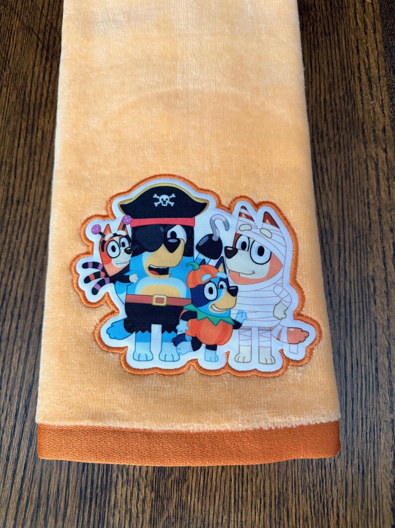 May include: A soft peach-colored hand towel featuring a central embroidered patch of four cartoon dogs dressed in Halloween costumes, including a pirate, a mummy, a pumpkin, and a spider, with a solid dark orange fabric border at the bottom edge.