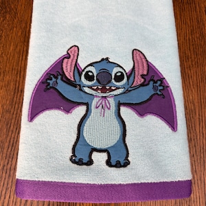 May include: A light blue hand towel featuring an embroidered graphic of the character Stitch with purple bat wings and a small purple bow tie. The towel has a solid purple fabric border along the bottom edge, providing a distinct color contrast.