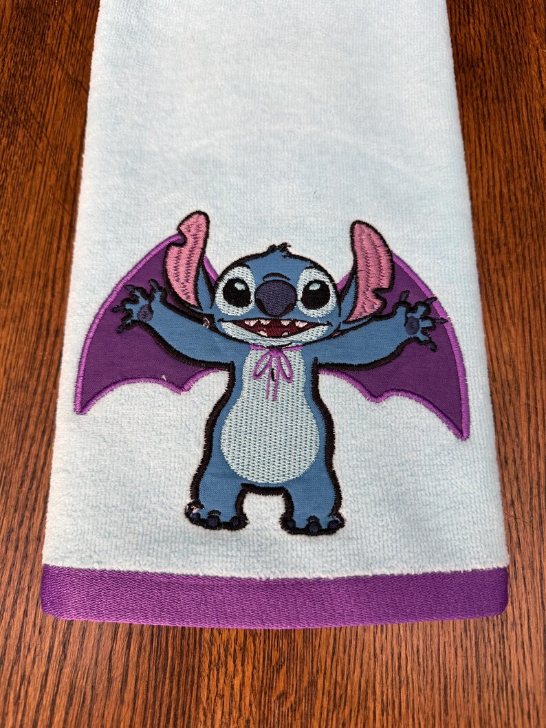 May include: A light blue hand towel featuring an embroidered graphic of the character Stitch with purple bat wings and a small purple bow tie. The towel has a solid purple fabric border along the bottom edge, providing a distinct color contrast.