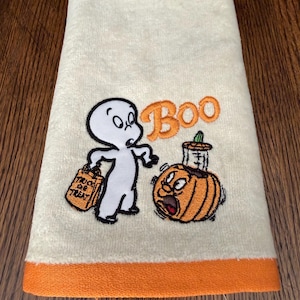May include: A cream-colored hand towel with an orange hem featuring an embroidered white ghost holding an orange trick-or-treat bag, an orange pumpkin with a surprised expression, and the orange embroidered text Boo above the pumpkin.