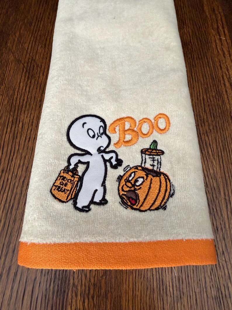May include: A cream-colored hand towel with an orange hem featuring an embroidered white ghost holding an orange trick-or-treat bag, an orange pumpkin with a surprised expression, and the orange embroidered text Boo above the pumpkin.