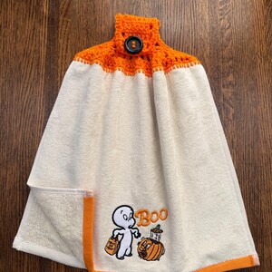 May include: A cream-colored hanging kitchen towel featuring an orange crocheted top with a black button closure. The towel displays an embroidered graphic of a ghost holding a trick-or-treat bag next to a pumpkin with the text Boo.