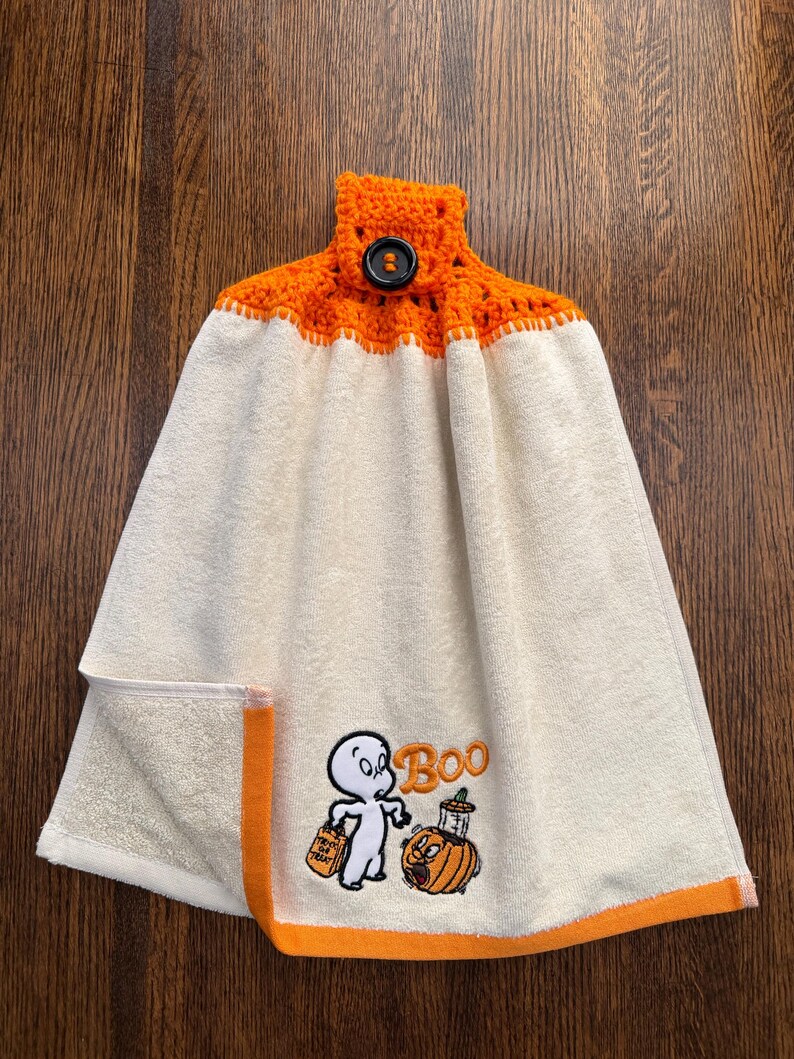 May include: A cream-colored hanging kitchen towel featuring an orange crocheted top with a black button closure. The towel displays an embroidered graphic of a ghost holding a trick-or-treat bag next to a pumpkin with the text Boo.