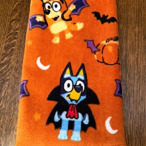 May include: An orange towel featuring a Halloween-themed pattern with cartoon dogs dressed as a vampire and a bat. The design includes black bats, orange pumpkins, and white crescent moons scattered across the fabric surface in a repeating print.