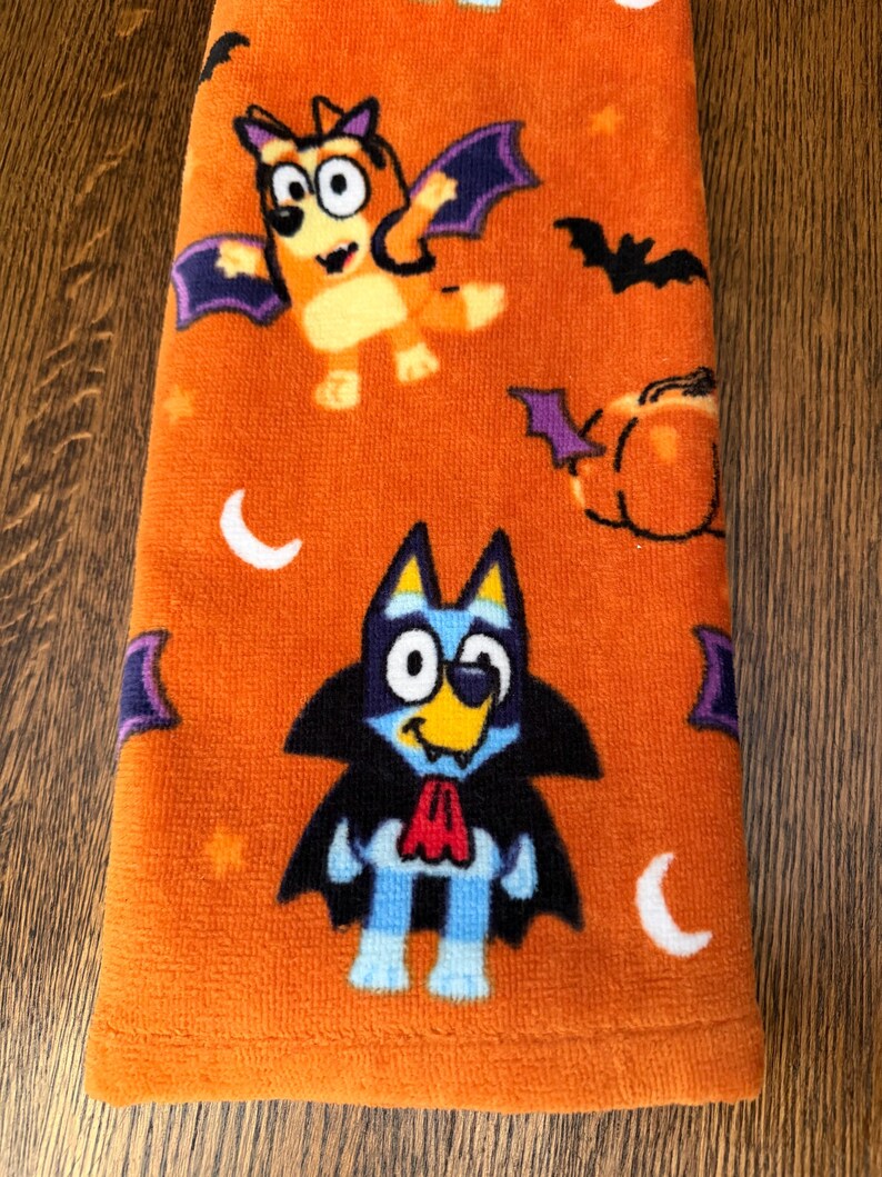 May include: An orange towel featuring a Halloween-themed pattern with cartoon dogs dressed as a vampire and a bat. The design includes black bats, orange pumpkins, and white crescent moons scattered across the fabric surface in a repeating print.