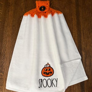 May include: A white hanging kitchen towel featuring a crocheted orange top with a black button closure. The towel displays an embroidered orange jack-o'-lantern with a small ghost on top and the word SPOOKY written in black lettering below it.