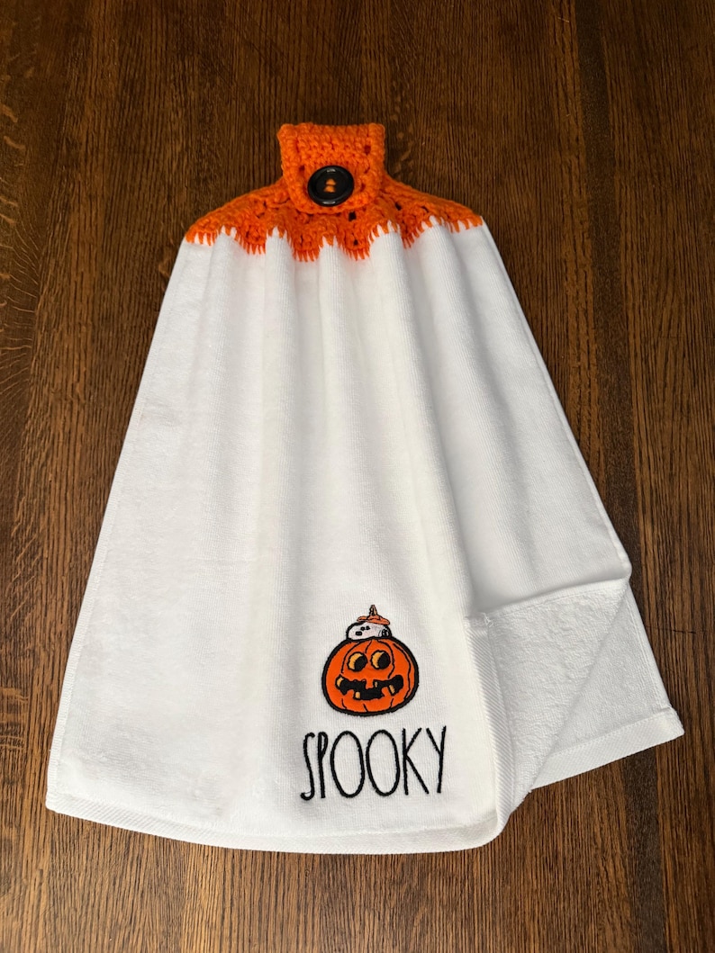 May include: A white hanging kitchen towel featuring a crocheted orange top with a black button closure. The towel displays an embroidered orange jack-o'-lantern with a small ghost on top and the word SPOOKY written in black lettering below it.