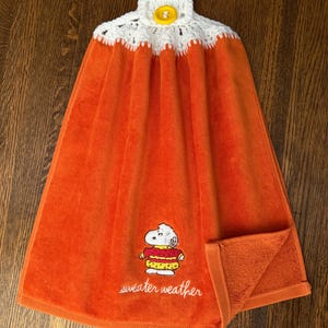 May include: An orange hanging kitchen towel featuring an embroidered Snoopy character wearing a red and yellow sweater with the text "sweater weather" below. The top has a white crocheted topper with a single yellow button for hanging.