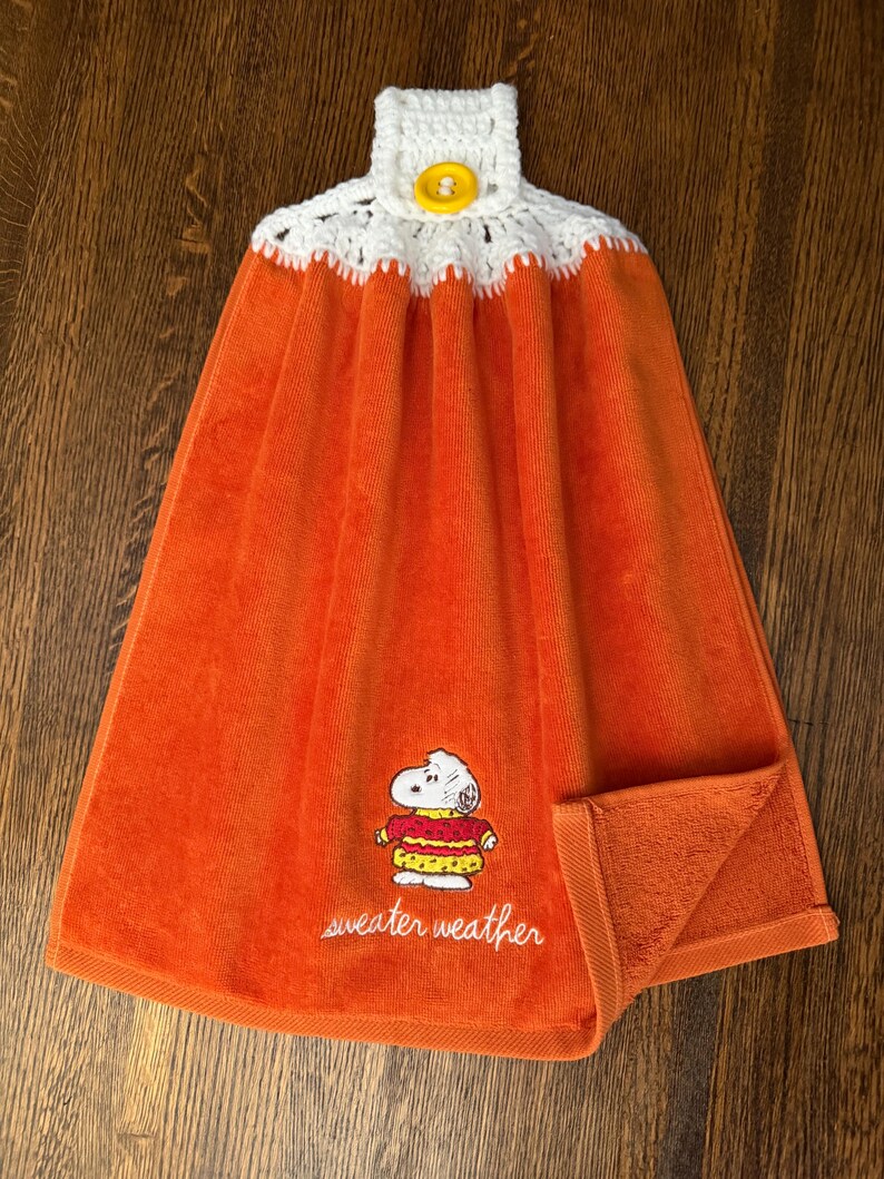 May include: An orange hanging kitchen towel featuring an embroidered Snoopy character wearing a red and yellow sweater with the text "sweater weather" below. The top has a white crocheted topper with a single yellow button for hanging.