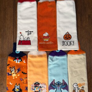 May include: Seven hanging kitchen towels with crocheted tops and buttons. Designs include Snoopy with text, a pumpkin with the word SPOOKY, Bluey characters in costumes, Stitch with wings, and a ghost with the word Boo on various colored fabric.