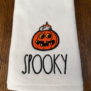 May include: A white hand towel featuring an embroidered orange jack-o'-lantern with a small white dog wearing an orange hat sitting on top. Below the pumpkin, the word SPOOKY is embroidered in black, stylized, handwritten-style lettering.