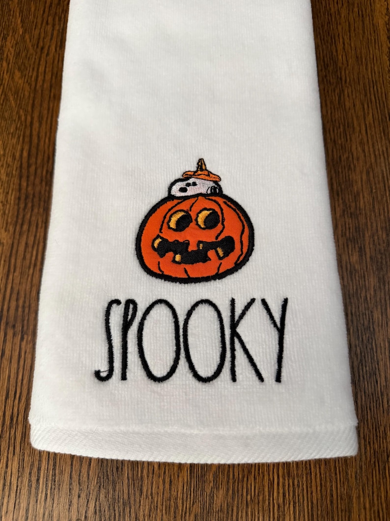 May include: A white hand towel featuring an embroidered orange jack-o'-lantern with a small white dog wearing an orange hat sitting on top. Below the pumpkin, the word SPOOKY is embroidered in black, stylized, handwritten-style lettering.