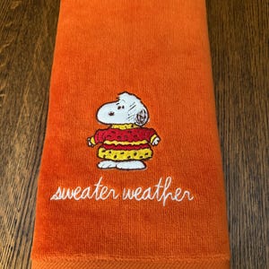 May include: A bright orange hand towel featuring an embroidered graphic of a white cartoon dog wearing a red and yellow patterned sweater. Below the character, the text "sweater weather" is embroidered in white cursive script lettering.