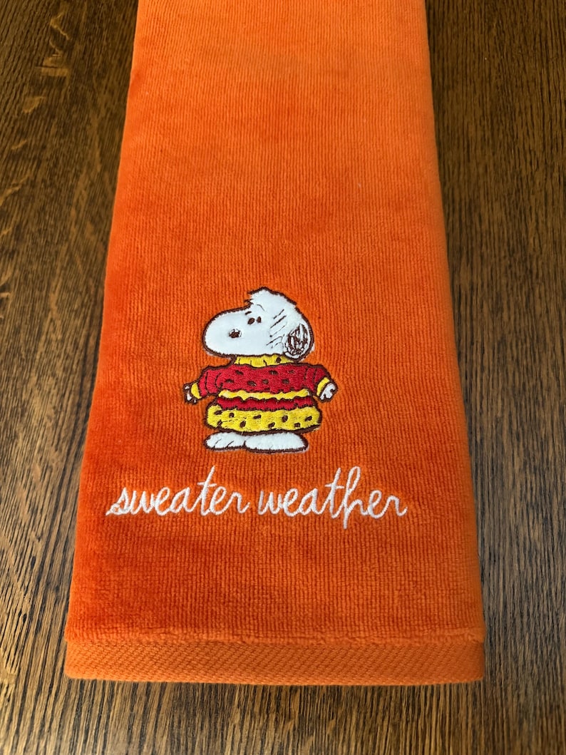 May include: A bright orange hand towel featuring an embroidered graphic of a white cartoon dog wearing a red and yellow patterned sweater. Below the character, the text "sweater weather" is embroidered in white cursive script lettering.