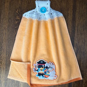 May include: A peach-colored hanging kitchen towel featuring a white crocheted top with a bright blue button. The towel displays a colorful embroidered patch of four cartoon dog characters dressed in various Halloween costumes including a pirate.
