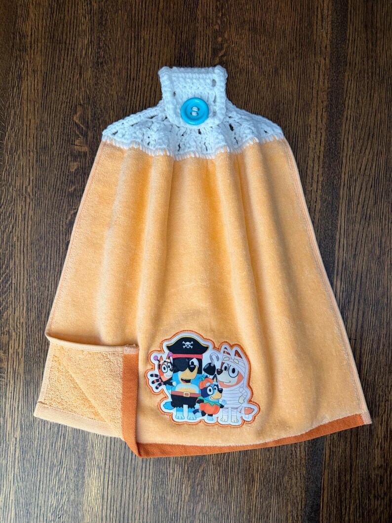May include: A peach-colored hanging kitchen towel featuring a white crocheted top with a bright blue button. The towel displays a colorful embroidered patch of four cartoon dog characters dressed in various Halloween costumes including a pirate.