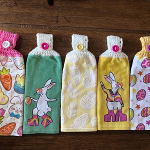 May include: Five Easter-themed kitchen towels with bunny and egg designs. The towels are white with different colored accents, including pink, yellow, green, and blue. Each towel has a crocheted border and a pink button.