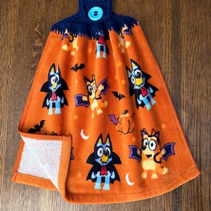 May include: An orange hanging kitchen towel featuring a repeating Halloween-themed pattern of cartoon dogs dressed as vampires and bats, accented with pumpkins and stars. The top has a dark blue crocheted topper secured with a light blue button.