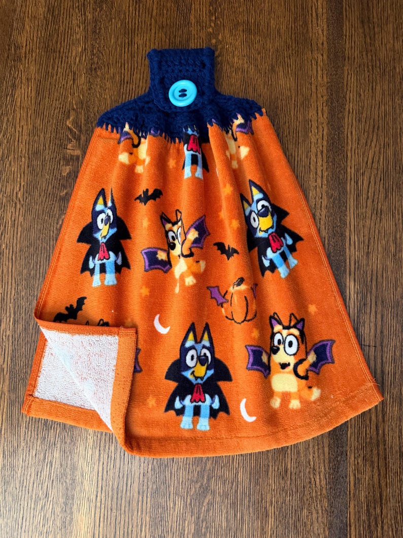 May include: An orange hanging kitchen towel featuring a repeating Halloween-themed pattern of cartoon dogs dressed as vampires and bats, accented with pumpkins and stars. The top has a dark blue crocheted topper secured with a light blue button.