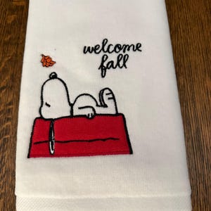 May include: A white kitchen towel featuring an embroidered design of a white dog lying on a red doghouse. Above the dog, the text reads "welcome fall" in black script, accompanied by a small, single orange embroidered autumn leaf.
