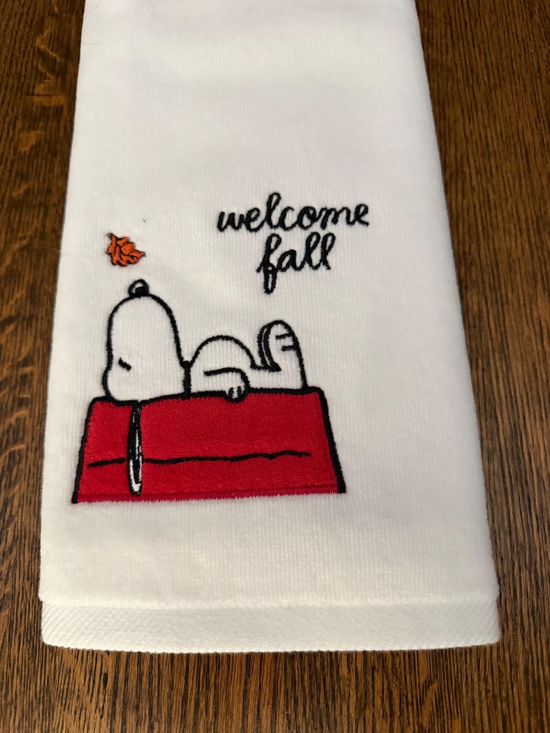 May include: A white kitchen towel featuring an embroidered design of a white dog lying on a red doghouse. Above the dog, the text reads "welcome fall" in black script, accompanied by a small, single orange embroidered autumn leaf.