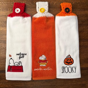 May include: Three hanging kitchen towels with crocheted tops and buttons. The left towel is white with Snoopy on a red doghouse and the text "welcome fall". The center is orange with Snoopy in a sweater and "sweater weather". The right is white with a pumpkin and "SPOOKY".