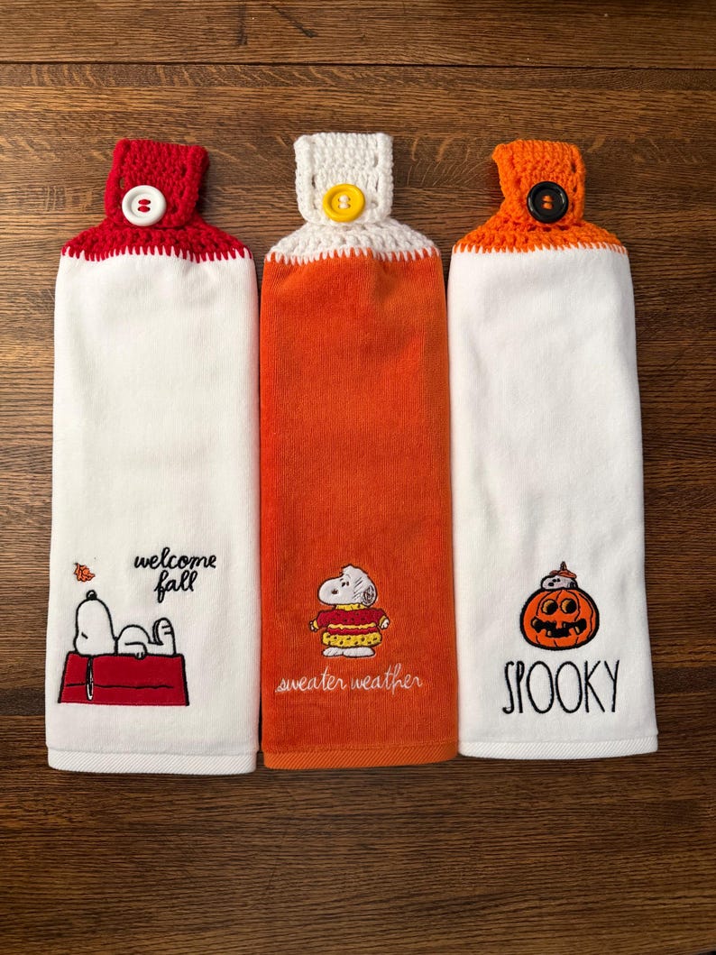 May include: Three hanging kitchen towels with crocheted tops and buttons. The left towel is white with Snoopy on a red doghouse and the text "welcome fall". The center is orange with Snoopy in a sweater and "sweater weather". The right is white with a pumpkin and "SPOOKY".