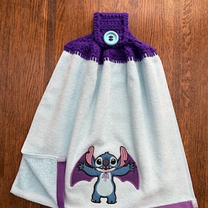 May include: A light blue hanging kitchen towel featuring a purple crocheted top with a light blue button. The towel displays an embroidered graphic of a blue alien character with large ears and purple wings, finished with a purple hem at the bottom.