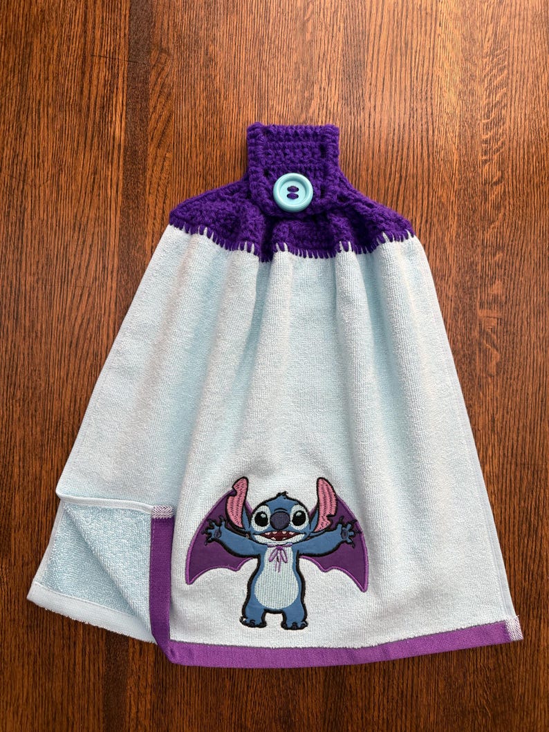 May include: A light blue hanging kitchen towel featuring a purple crocheted top with a light blue button. The towel displays an embroidered graphic of a blue alien character with large ears and purple wings, finished with a purple hem at the bottom.
