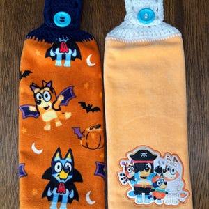 May include: Two hanging kitchen towels with crocheted tops and bright blue buttons. The left towel is orange with a pattern of cartoon dogs dressed as vampires, bats, and pumpkins. The right towel is solid light orange with a patch of cartoon dogs in costumes.