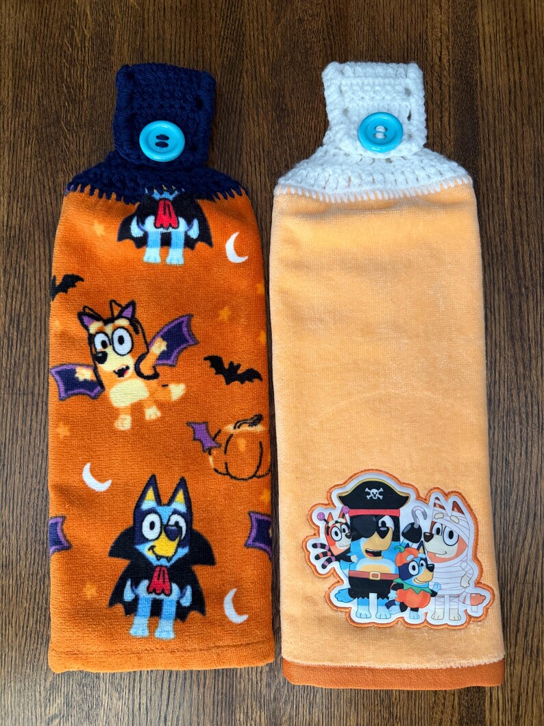 May include: Two hanging kitchen towels with crocheted tops and bright blue buttons. The left towel is orange with a pattern of cartoon dogs dressed as vampires, bats, and pumpkins. The right towel is solid light orange with a patch of cartoon dogs in costumes.