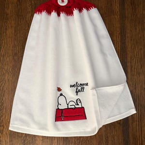 May include: A white hanging kitchen towel with a red crocheted top featuring a white button. The towel displays an embroidered graphic of a white dog lying on a red doghouse with the text "welcome fall" and a small orange leaf above it.