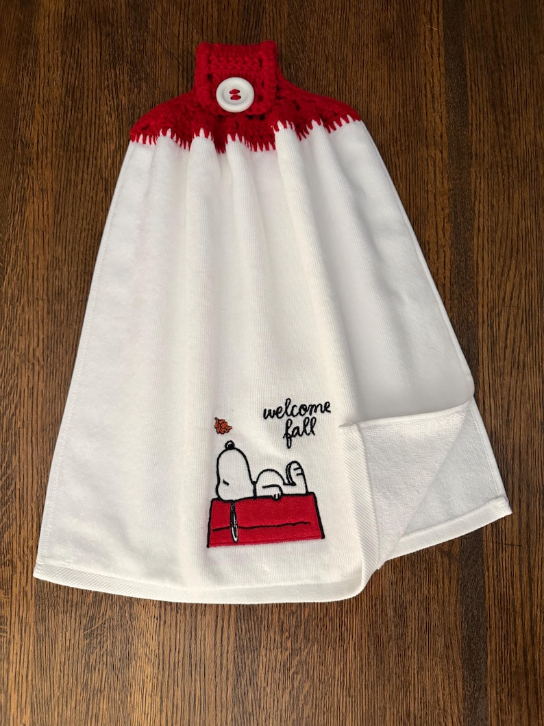 May include: A white hanging kitchen towel with a red crocheted top featuring a white button. The towel displays an embroidered graphic of a white dog lying on a red doghouse with the text "welcome fall" and a small orange leaf above it.