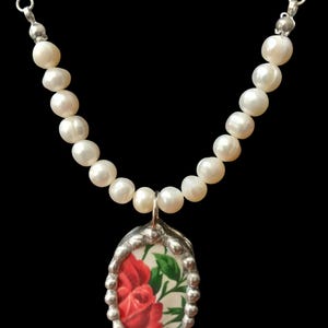 May include: A pearl necklace with a silver pendant. The pendant features a red rose design on a white background, framed by a silver border. The necklace has a black background.