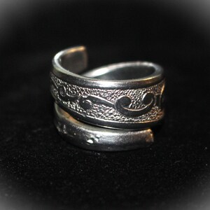 May include: A silver ring with a swirling, embossed design. The ring is made of two bands that are twisted together.