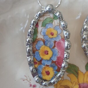 May include: Oval pendant necklace with a floral design. The pendant features a silver-colored beaded frame surrounding a ceramic insert with blue, yellow, and red flowers on a white background. The necklace has a silver chain.