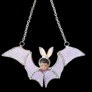 Könnte beinhalten: Ein Fledermaus-Sonnenfänger aus Glas mit einer Kinderfigur mit Hasenohren. Die lavendelfarbenen Flügel haben rosa Herzen und Wirbel, die mit Silber umrandet sind. Eine silberne Kette ist zum Aufhängen befestigt.