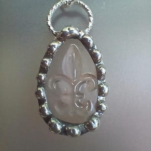 May include: A silver pendant necklace featuring a teardrop-shaped, light pink gemstone with a fleur-de-lis design. The gemstone is encased in a silver bezel with small, round accents. The chain is also silver.