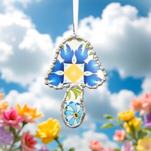 Puede incluir: Un adorno de cerámica de hongo azul y blanco con ribete plateado. El adorno está colgado de una cinta blanca sobre un fondo de cielo azul.