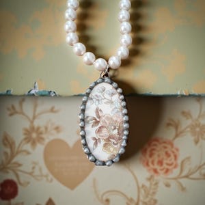 May include: A pearl necklace with a floral pendant. The oval pendant has a silver-colored border and features a cream-colored background with brown, tan, and blue floral designs. The necklace is displayed against a floral wallpaper background.