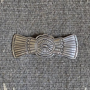 May include: A silver-toned metal belt buckle with an intricate design. The buckle features a central oval shape with radiating lines, flanked by fan-shaped elements. The background is a gray and cream-colored patterned textile.
